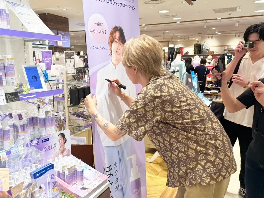 ファン必見!パク・ユチョン直筆サイン入り等身大バナーが当たる!AROMA BLOOMブラックフライデーで唯一無二のチャンス 2 ある男性が、パク・ユチョンのポスターにサインをしている様子を写した写真です。周りには、商品が陳列されている店内の様子が写っています。
