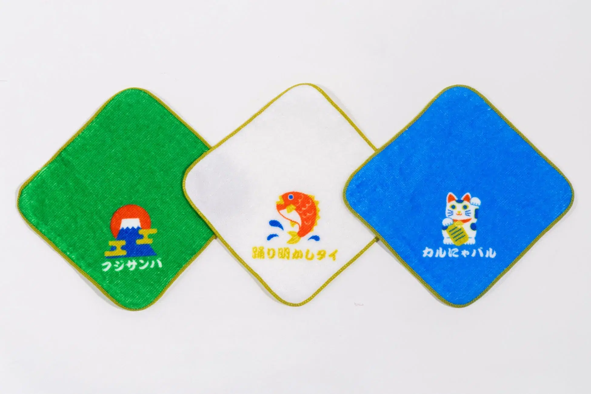 3つのハンカチが並んでいる写真。それぞれに異なるイラストと日本語の文字が描かれている。左は富士山と太陽のイラスト、中央は魚のイラスト、右は招き猫のイラスト。
