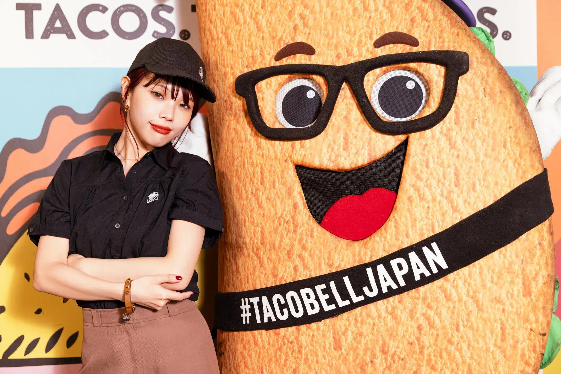 タコスベルのキャラクターと女性が写っている写真です。女性はタコスベルの制服を着ており、キャラクターの隣でポーズをとっている.