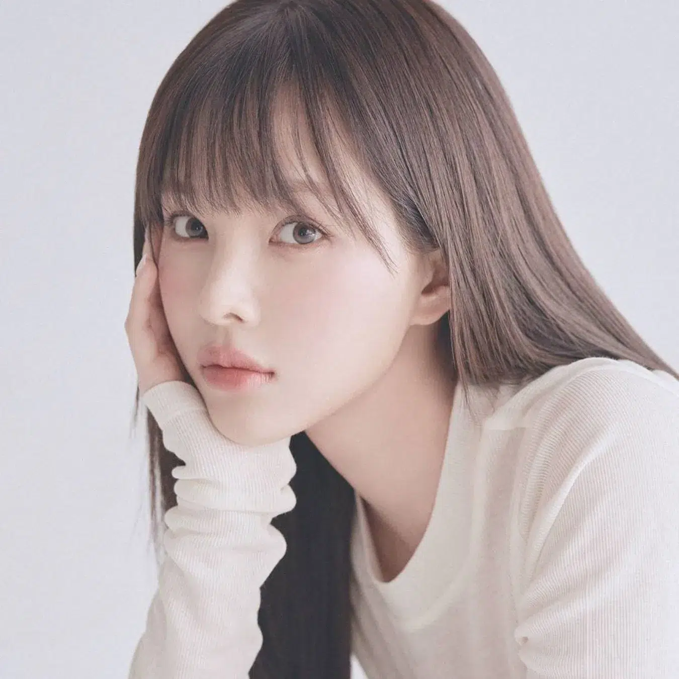 LE SSERAFIM専属メイクアップアーティスト監修!Pyt「GENBA LIP GLOSS」から待望のホリデー限定2色が登場 1 女性のポートレート。長い茶色の髪で、白い服を着て、顔を手に当てている。