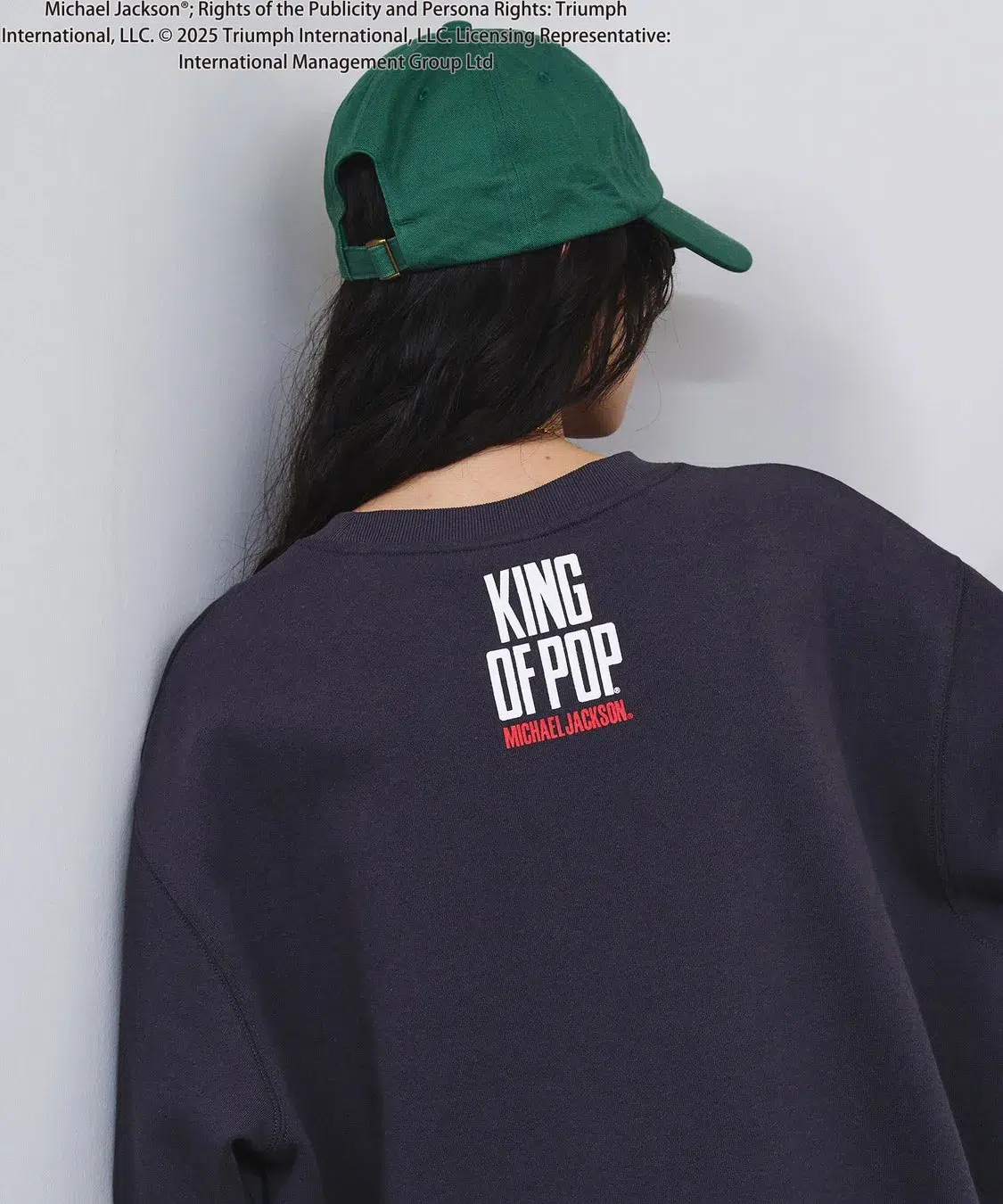 「KING OF POP」を纏う!ROSE BUDがマイケル・ジャクソン特別コレクションを初発表、ジェンダーレスで楽しめる全貌を公開! 3 後ろ姿の女性が、マイケル・ジャクソンの「KING OF POP」と書かれたトレーナーを着て、緑色の帽子をかぶっている画像です。