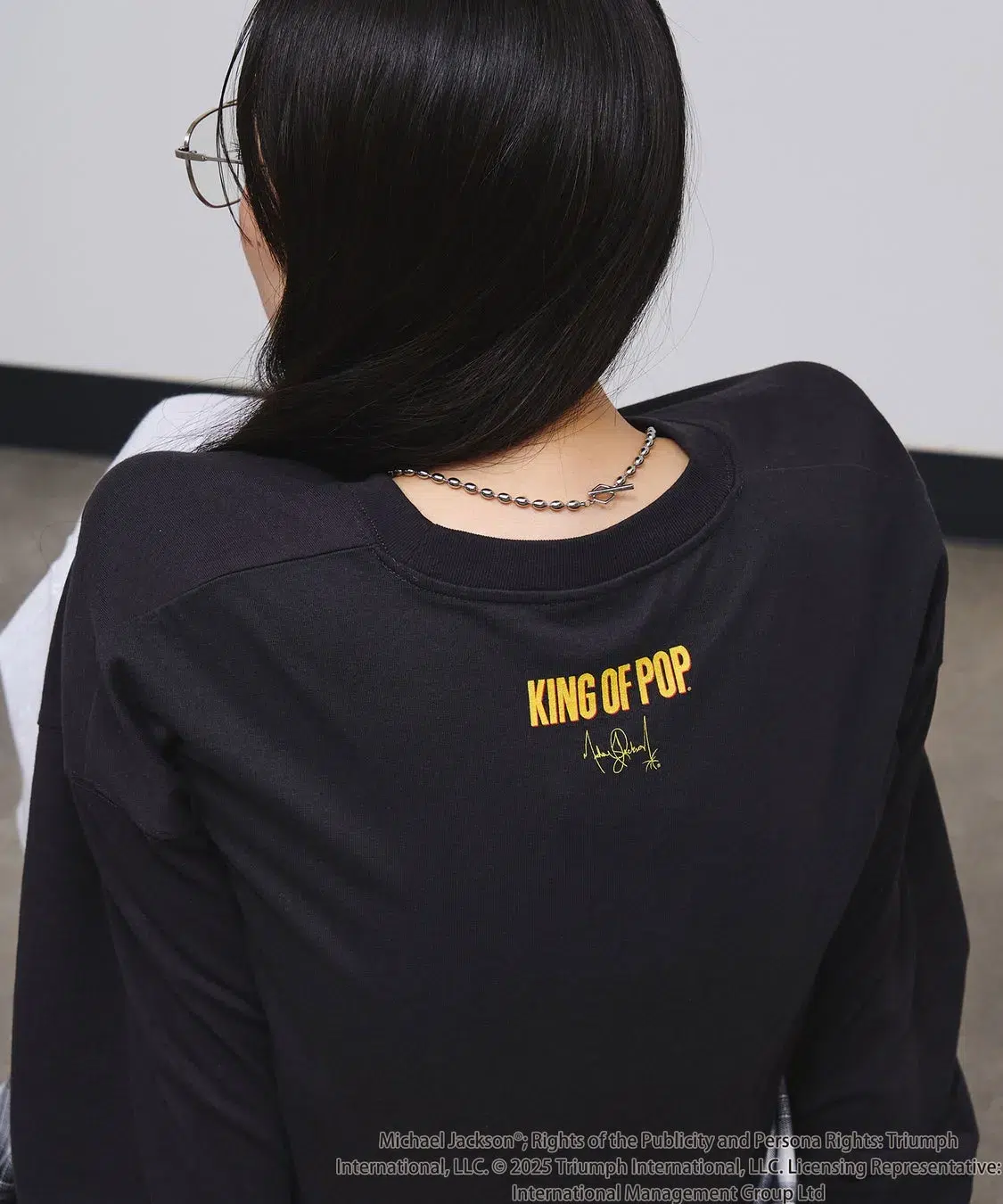 「KING OF POP」を纏う!ROSE BUDがマイケル・ジャクソン特別コレクションを初発表、ジェンダーレスで楽しめる全貌を公開! 7 後ろ姿の女性が写っており、黒い長袖の服を着ています。服には「KING OF POP」という文字とサインがプリントされています。