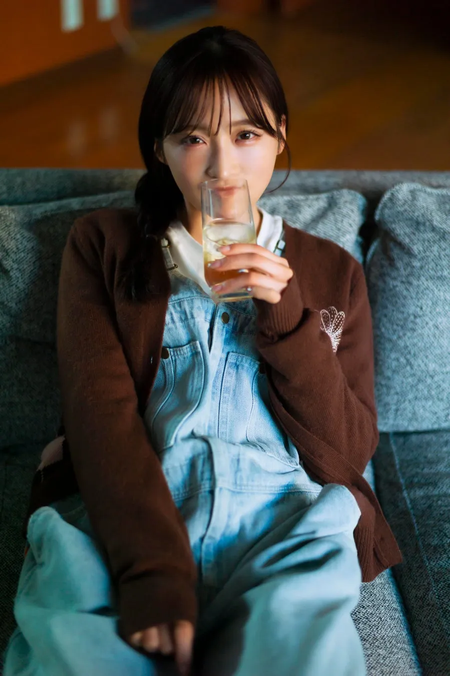 デニムのオーバーオールと茶色のカーディガンを着た小栗有以さんがソファに座り、グラスに入った飲み物を飲んでいる。