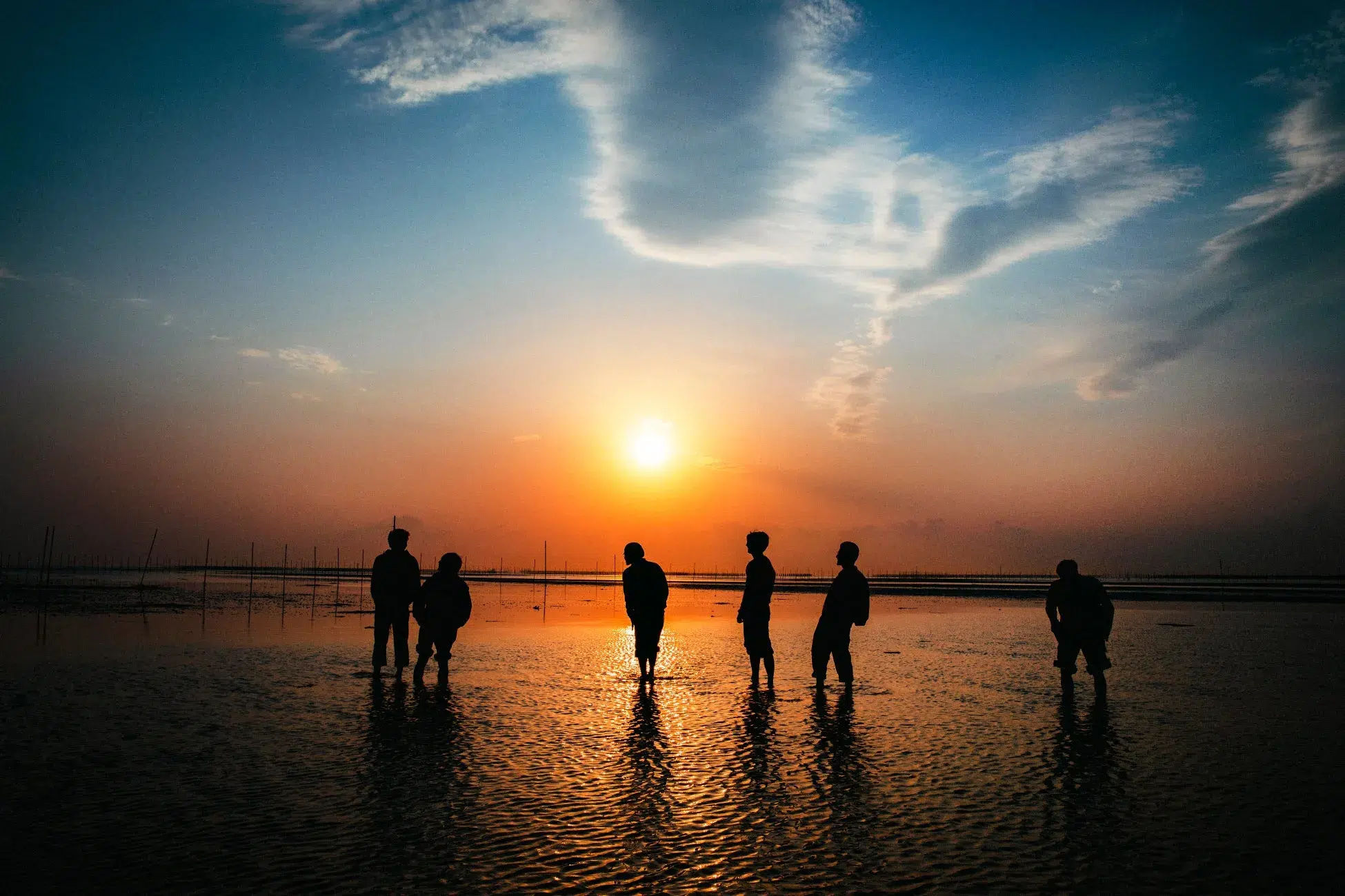 夕焼け空の下、水面に人が立っているシルエットを捉えた写真。オレンジ色の夕日が水面に反射し、美しい光景を作り出している。人々は水遊びをしているようにも見える。