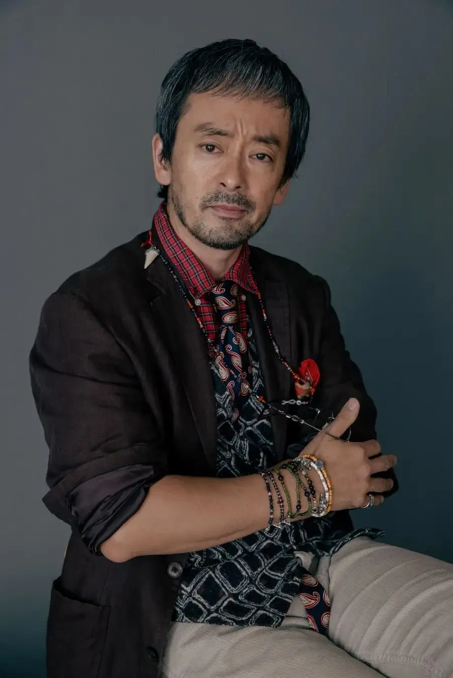 男性が椅子に座って腕を組んでいるポートレート写真。彼は茶色のジャケット、チェックのシャツ、ネクタイ、ブレスレットを着用しています。彼の顔にはひげとわずかに笑った表情があります。