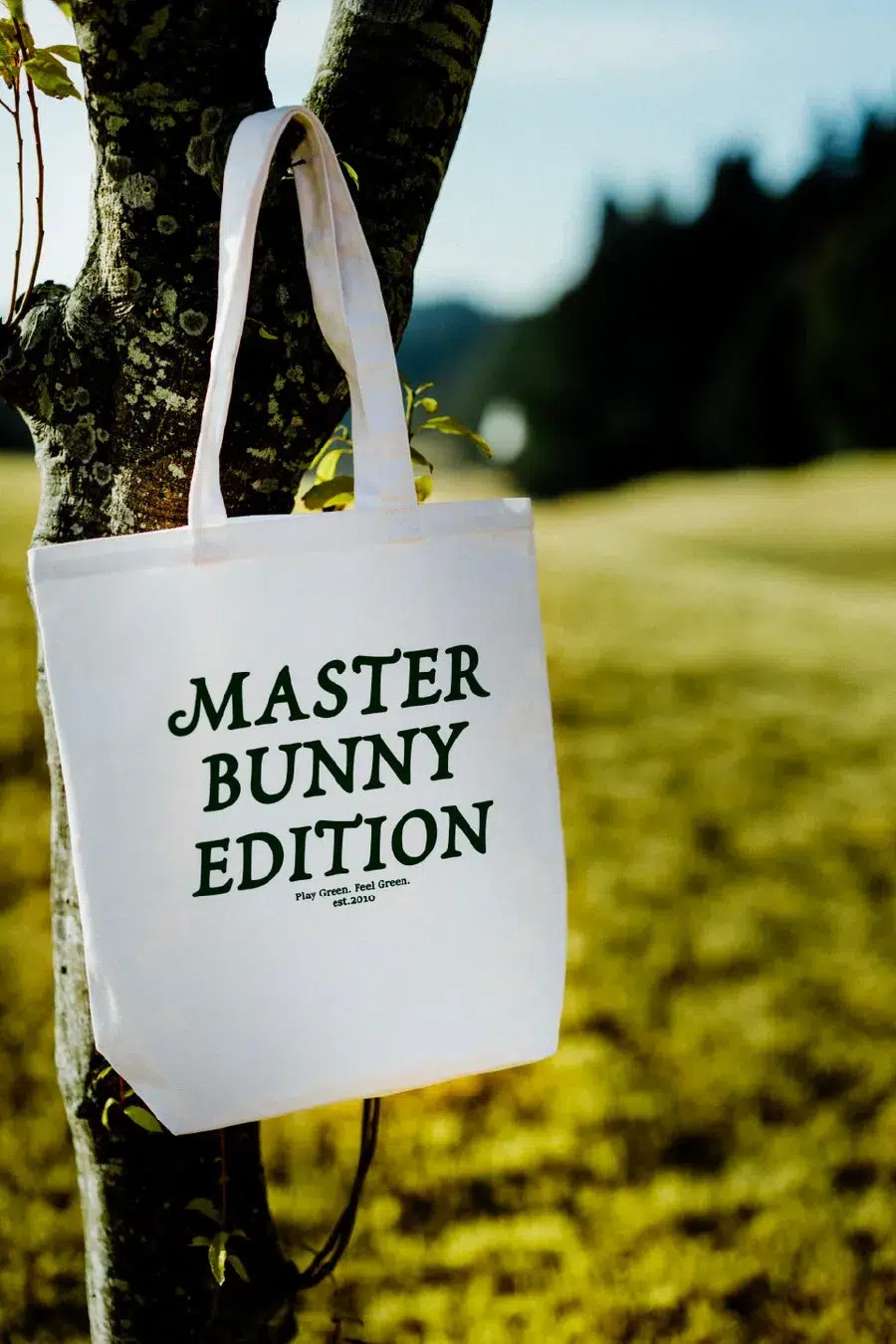 木にかけられた白いトートバッグ。バッグには「MASTER BUNNY EDITION」の文字と「Play Green. Feel Green. est.2010」の文字が緑色で書かれている。背景には緑色の芝生と木々が写っている。