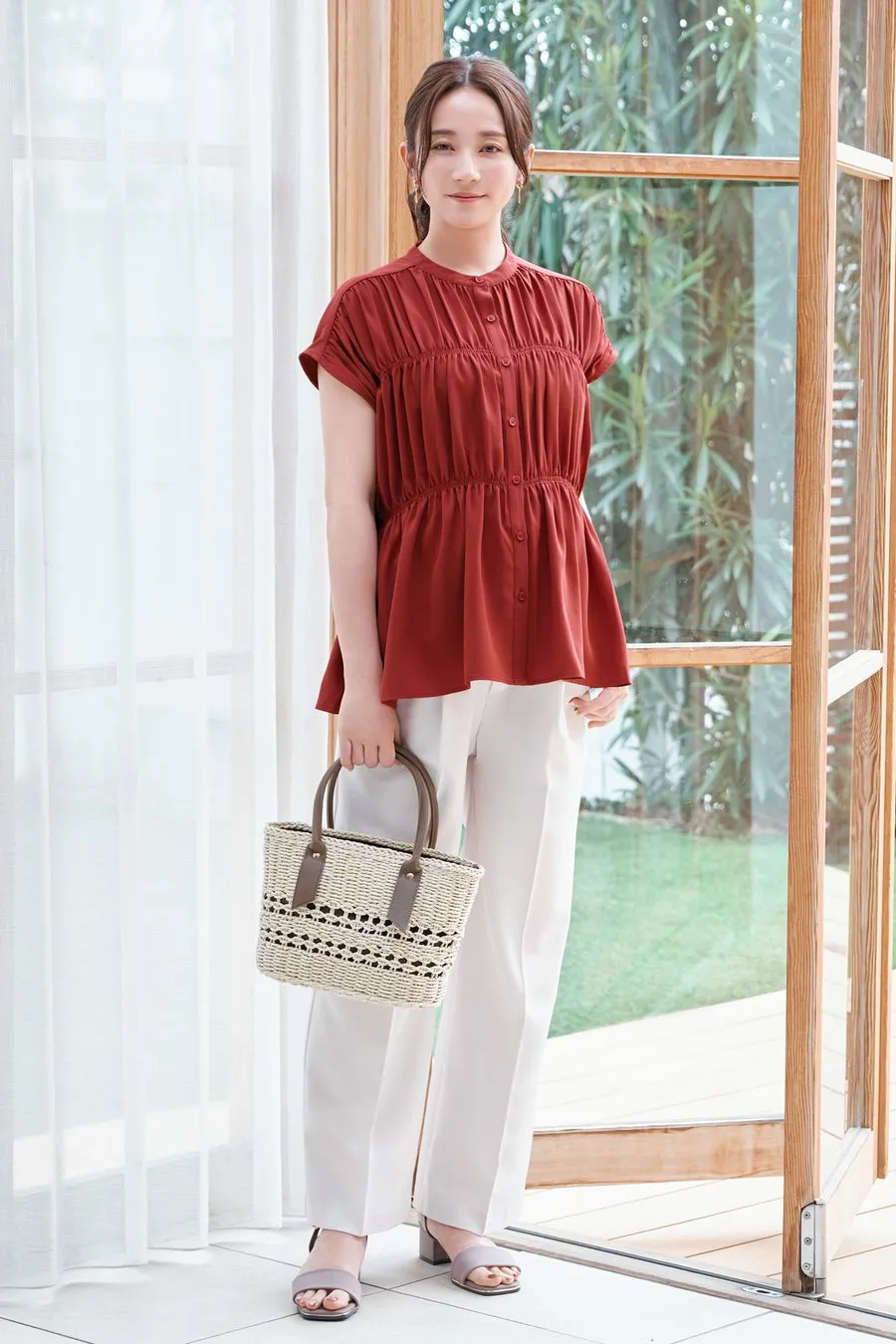 女性が、赤いトップスと白いパンツを着て、カゴバッグを持ち、窓の前に立っている写真です。