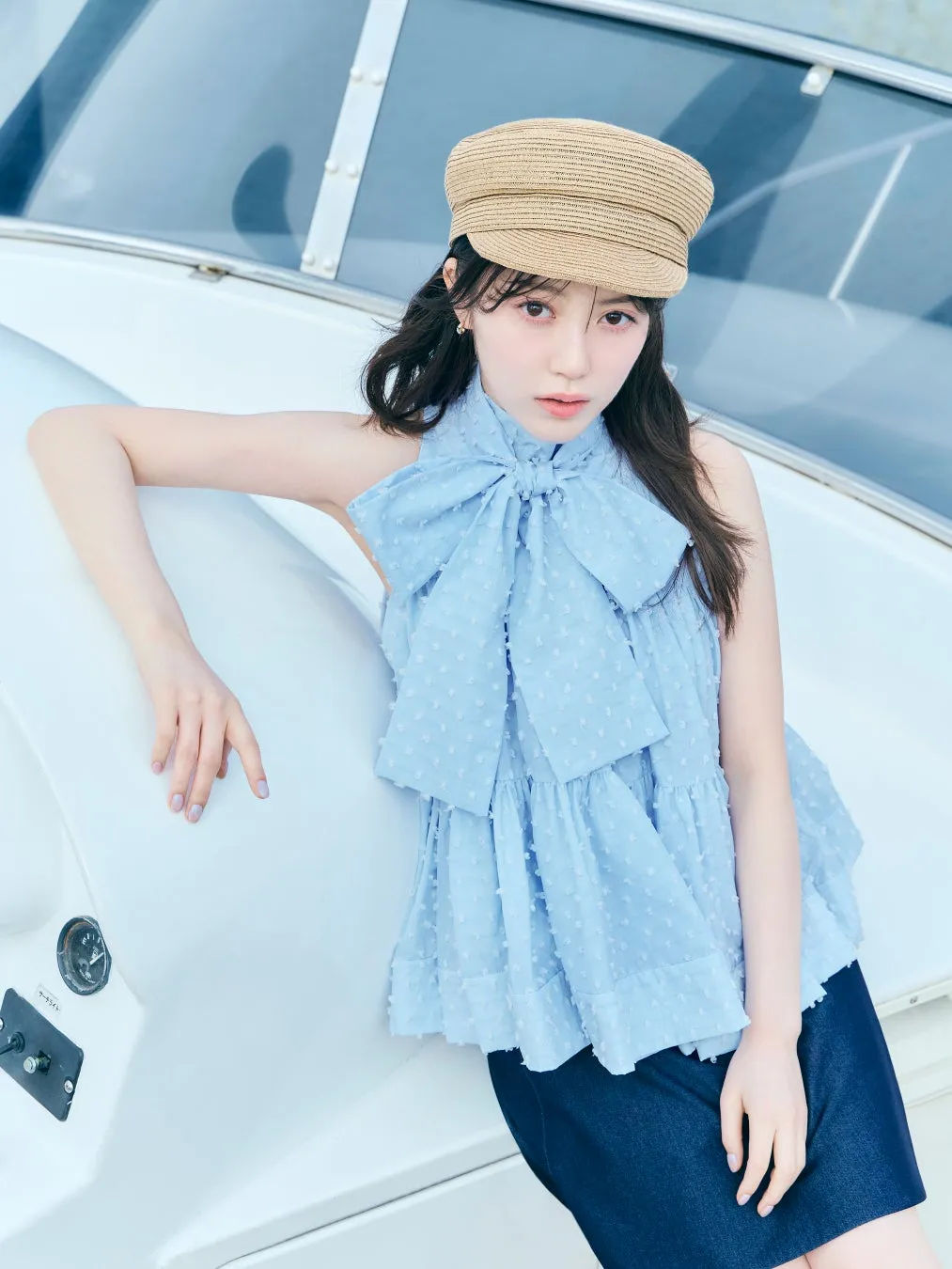 女性がヨットの上でポーズを取っている写真。彼女は水色のトップスとジーンズを着用し、帽子をかぶっています。