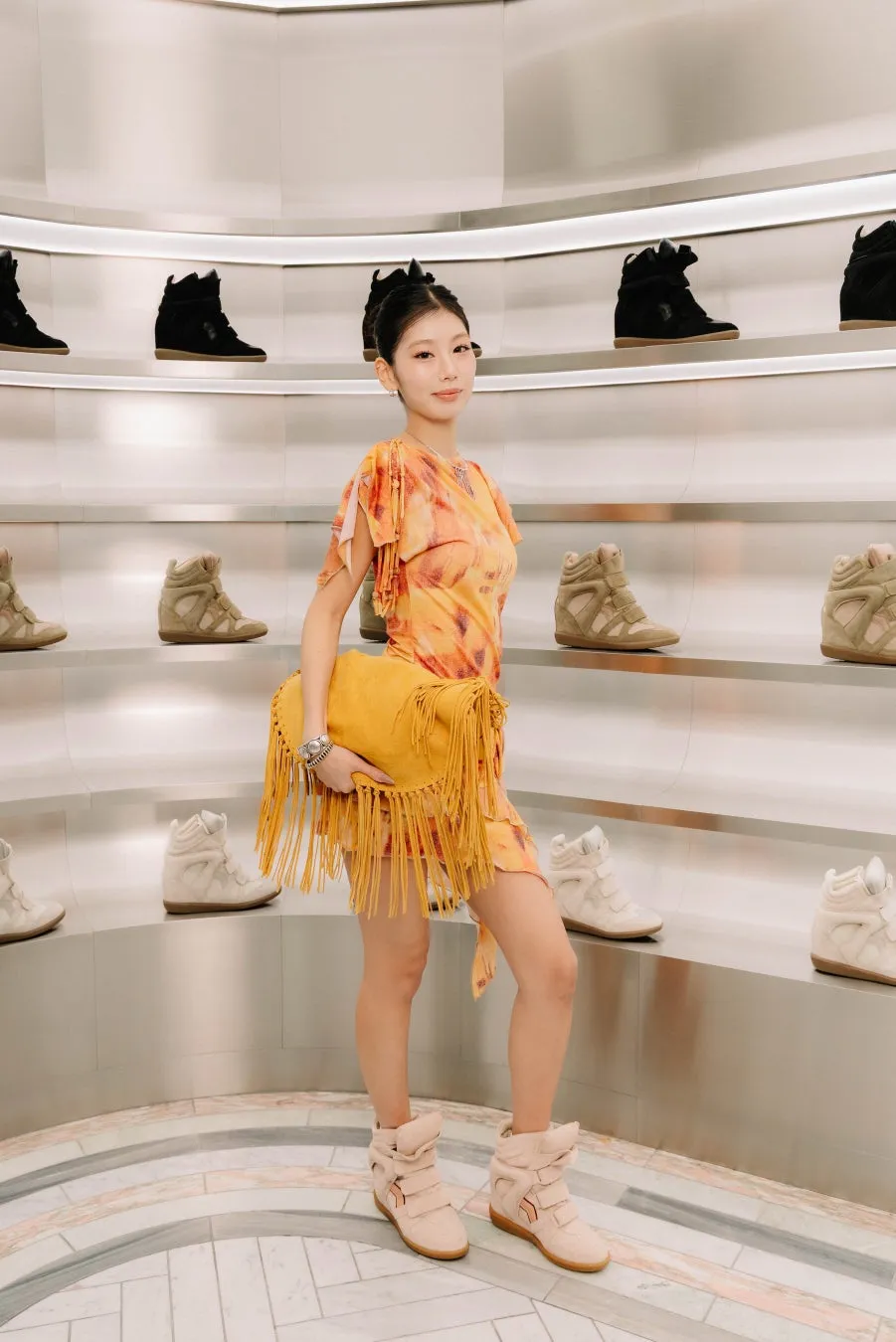 女性が黄色いフリンジバッグを持ち、オレンジ色のミニドレスを着て、靴が並んだ棚の前で立っている写真です。
