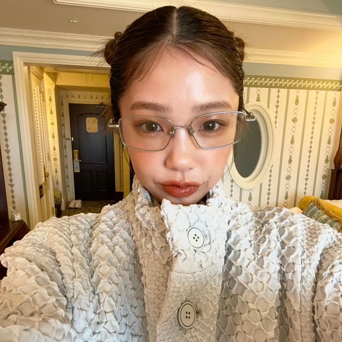 眼鏡をかけた女性の自撮り写真。白い波状のジャケットを着ており、背景にはホテルの部屋と思われる場所が写っています。