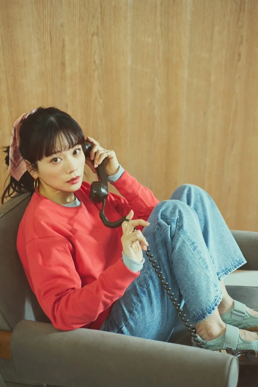 女性が電話を持ってソファに座っている写真。女性は赤いセーターとジーンズを着ており、ピンクのスカーフを頭に巻いている。背景は木目調。