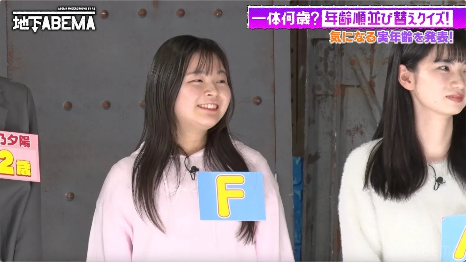 女性が出演している日本のテレビ番組のワンシーン。出演者は、ピンクの服を着た女性と、白いセーターを着た女性の2名。女性たちの前には、それぞれ「F」と書かれた札と、年齢を示唆する札がある。番組のロゴやタイトルも確認できる。