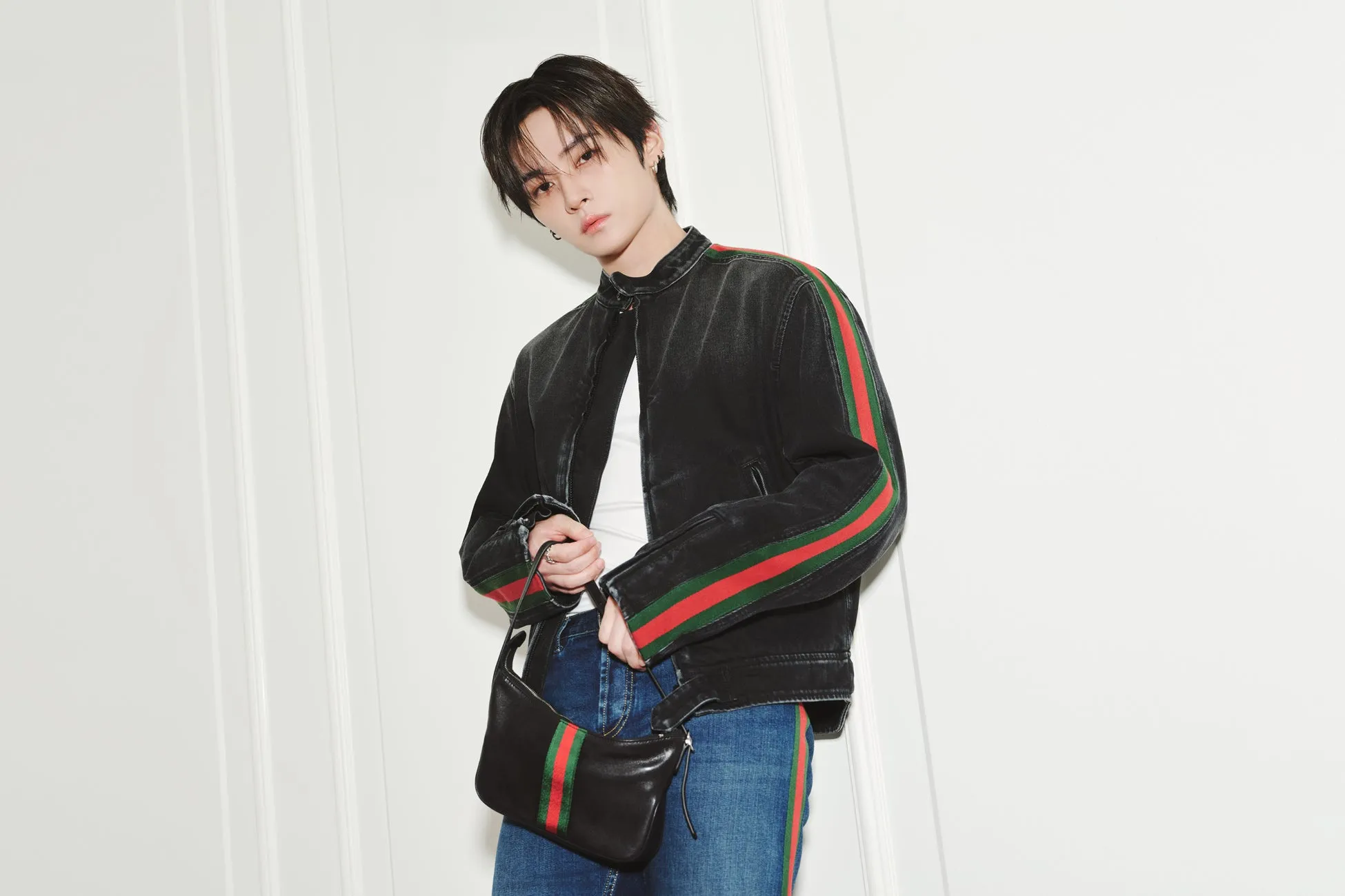 Stray Kidsリノがグッチ「Generation Gucci」で魅せた新境地!唯一無二の洗練ルック2選を徹底解剖 2 黒いジャケットとバッグを持った男性が立っている写真です。ジャケットには赤と緑のストライプが入っています。男性は白いシャツと青いジーンズを着ています。